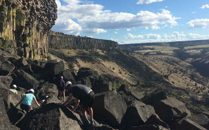a group of students make their way along a rocky landscape to a rock wall for climbing